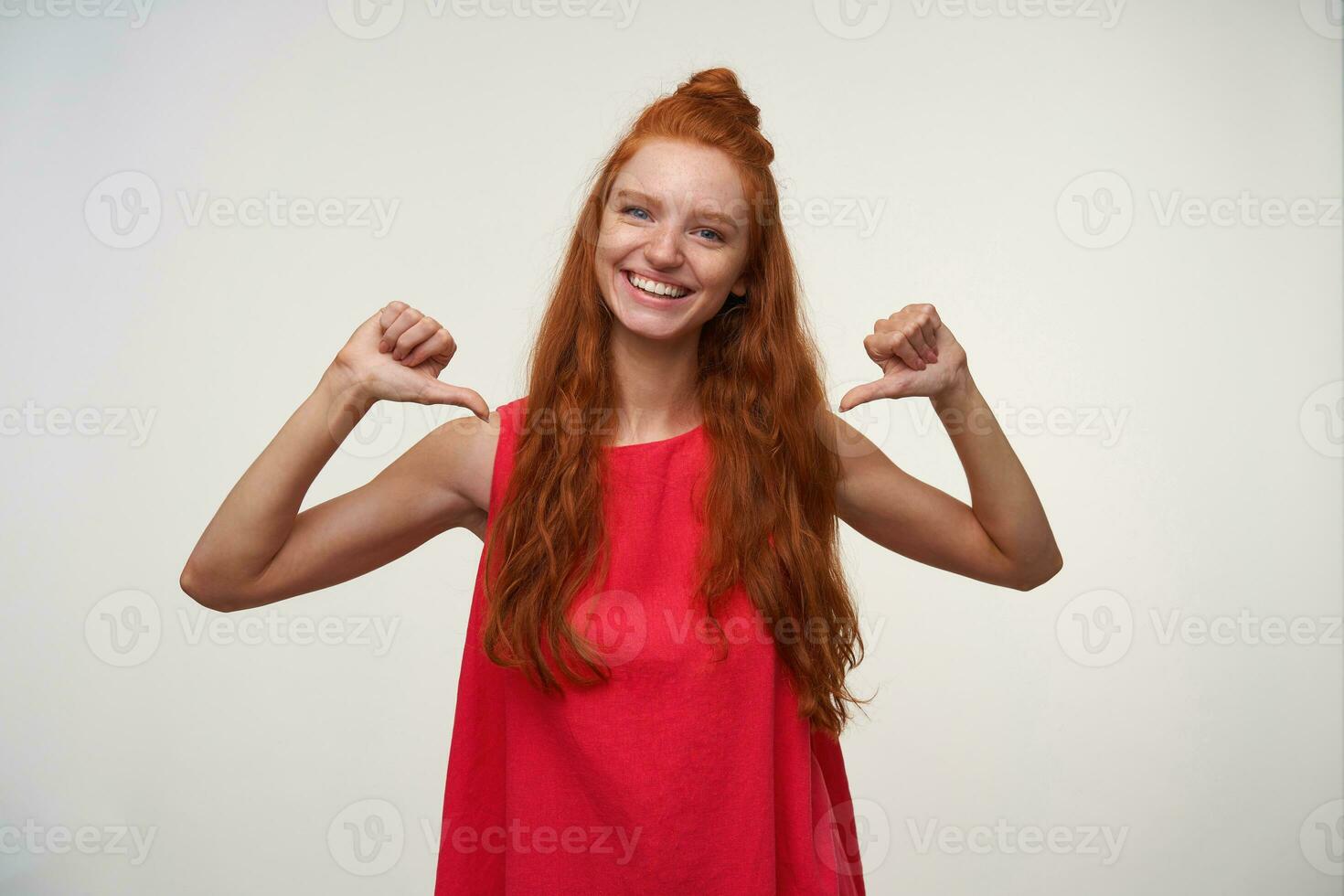 Portrait of happy young woman with foxy wavy hair wearing casual pink dress, posing over white background without make up, pointing to herself with thumbs and smiling broadly to camera photo