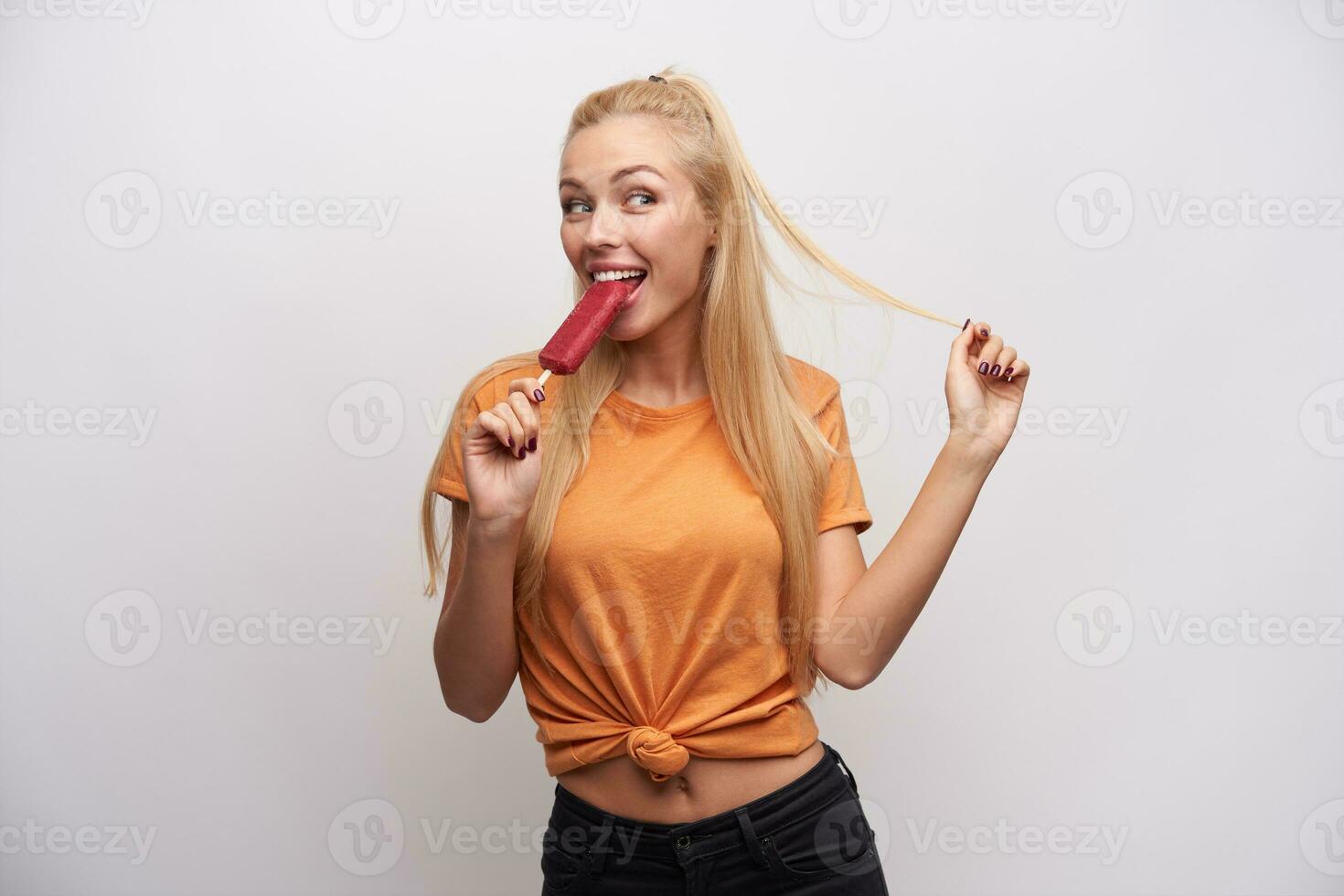 alegre joven hermosa hembra tracción su largo rubia pelo con mano mientras comiendo helado en palo, mirando felizmente aparte y sonriente atentamente, en pie en contra blanco antecedentes foto