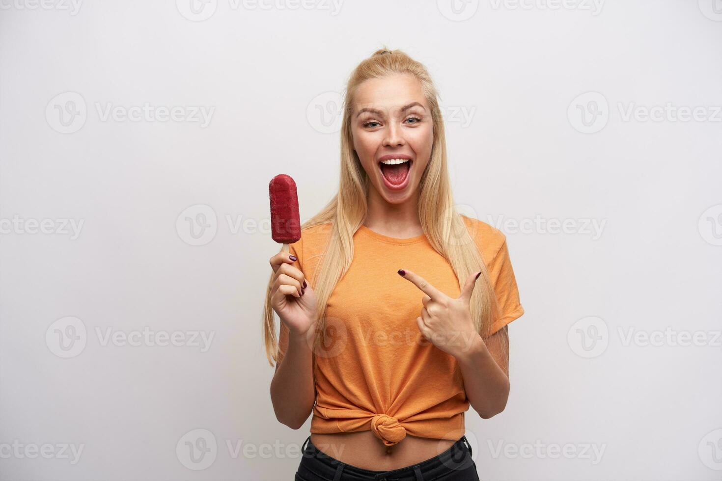 Overjoyed lovely young blonde woman with casual hairstyle pointing with forefinger on berry ice-cream in her hand, looking joyfully to camera with wide mouth opened, isolated over white background photo