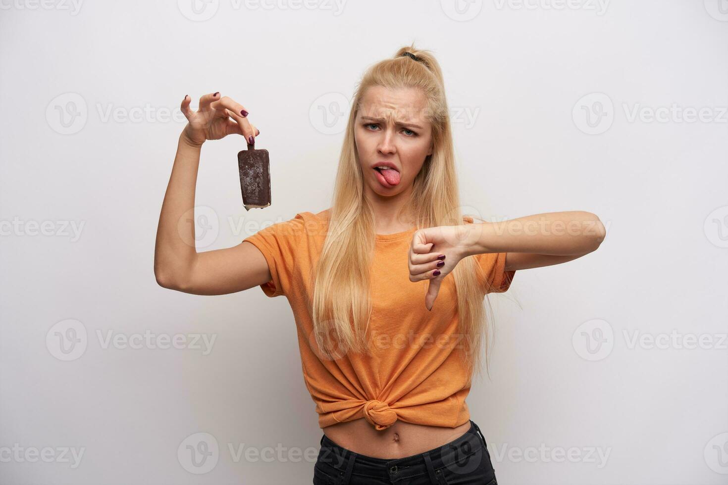 Displeased young long haired blonde female with ponytail hairstyle frowning her face and sticking out tongue, holding ice-cream and showing down with thumb, isolated over white background photo