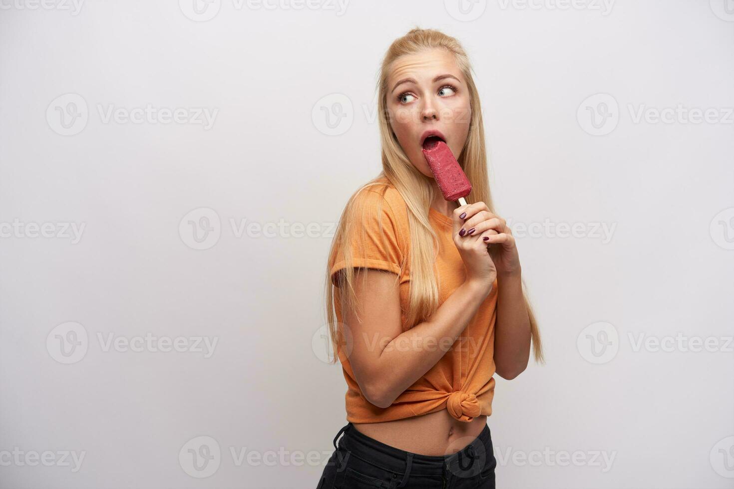 Confused attractive young long haired blonde lady in casual clothes looking over her shoulder and wrinkling forehead, keeping berry ice-cream while posing over white background photo