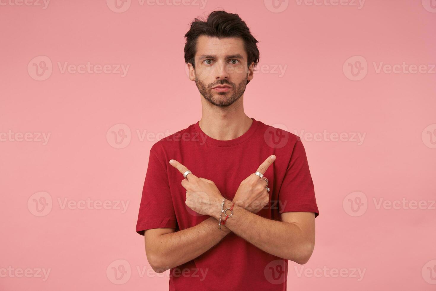 Studio photo of dark haired male with trendy haircut posing over pink background in red t-shirt, looking to camera with calm face and folded lips, showing with forefingers in different sides