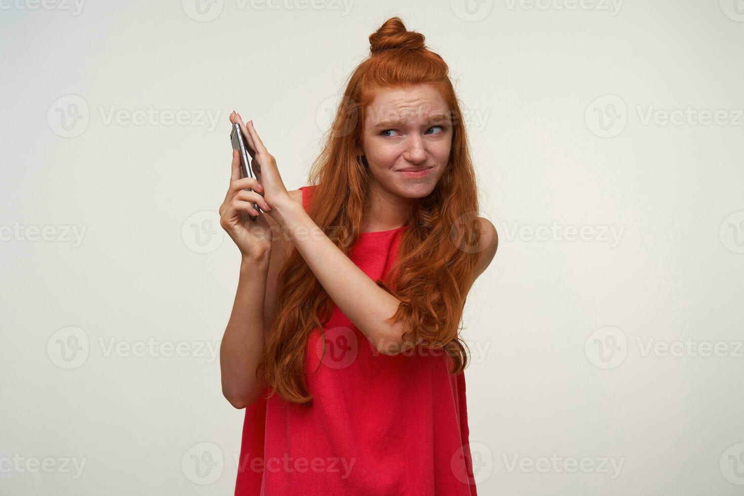 interior Disparo de joven cabeza lectora hembra vistiendo su pelo en nudo, posando terminado blanco antecedentes en casual vestido, teniendo desagradable conversacion con su teléfono inteligente y cubierta auricular a evitar sonidos foto