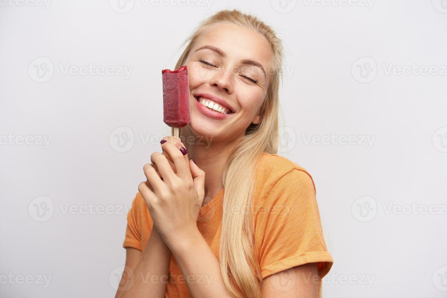 Pleased good looking pretty young blonde female in casual clothes holding ice-cream on stick and smiling sincerely with closed eyes, standing against white background photo