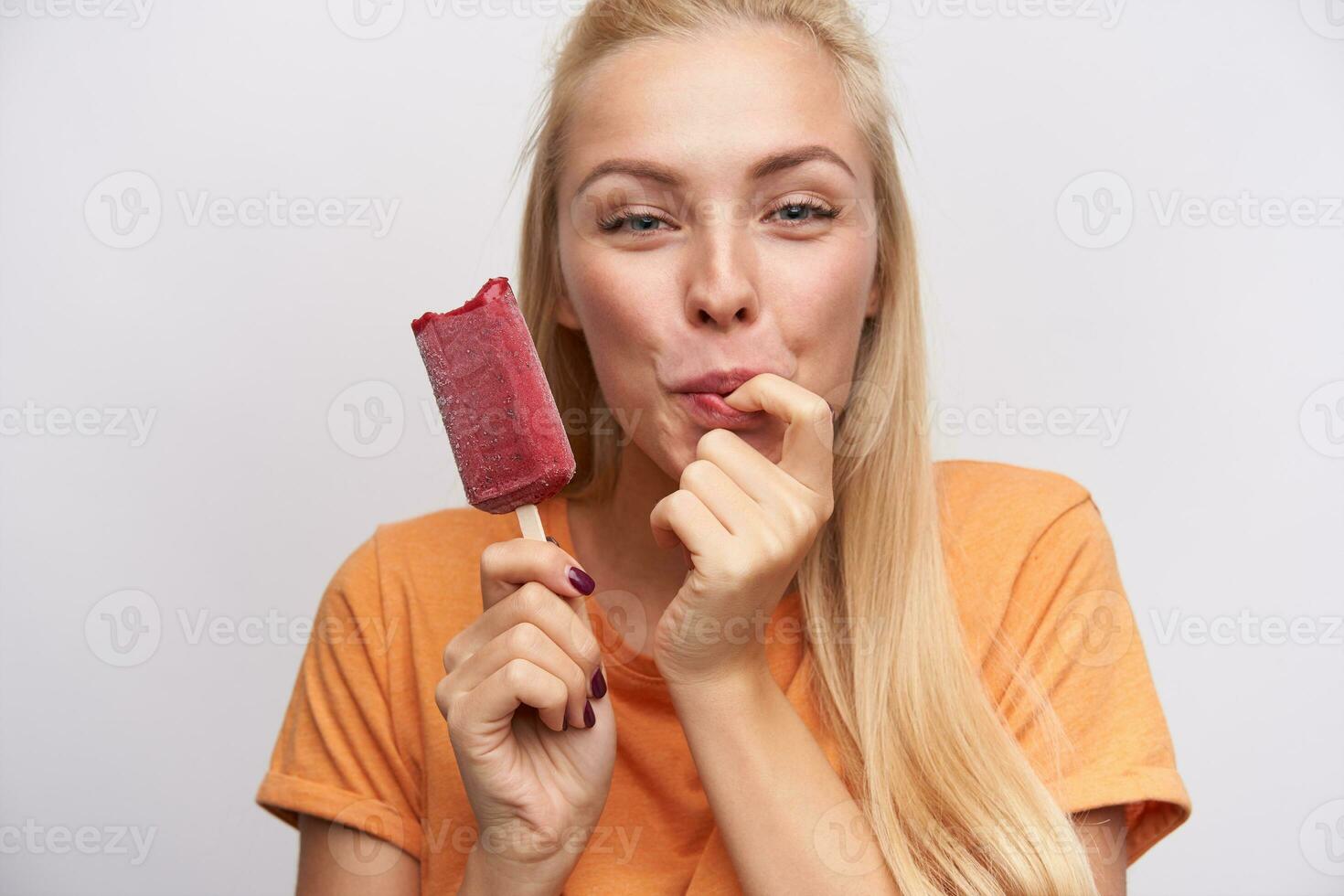 Close-up of positive lovely young long haired blonde lady delighting berry ice-cream while posing over white background, licking her fingers and looking cheerfully at camera photo