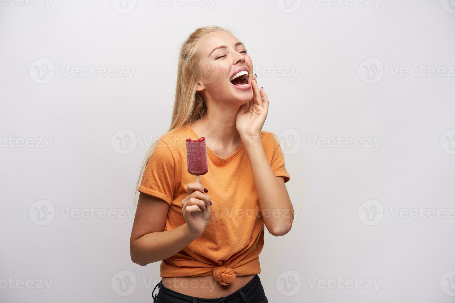 Studio shot of pleasant looking positive young blonde female with ice-cream in raised hand touching gently her face and laughing happily with closed eyes, isolated over white background photo