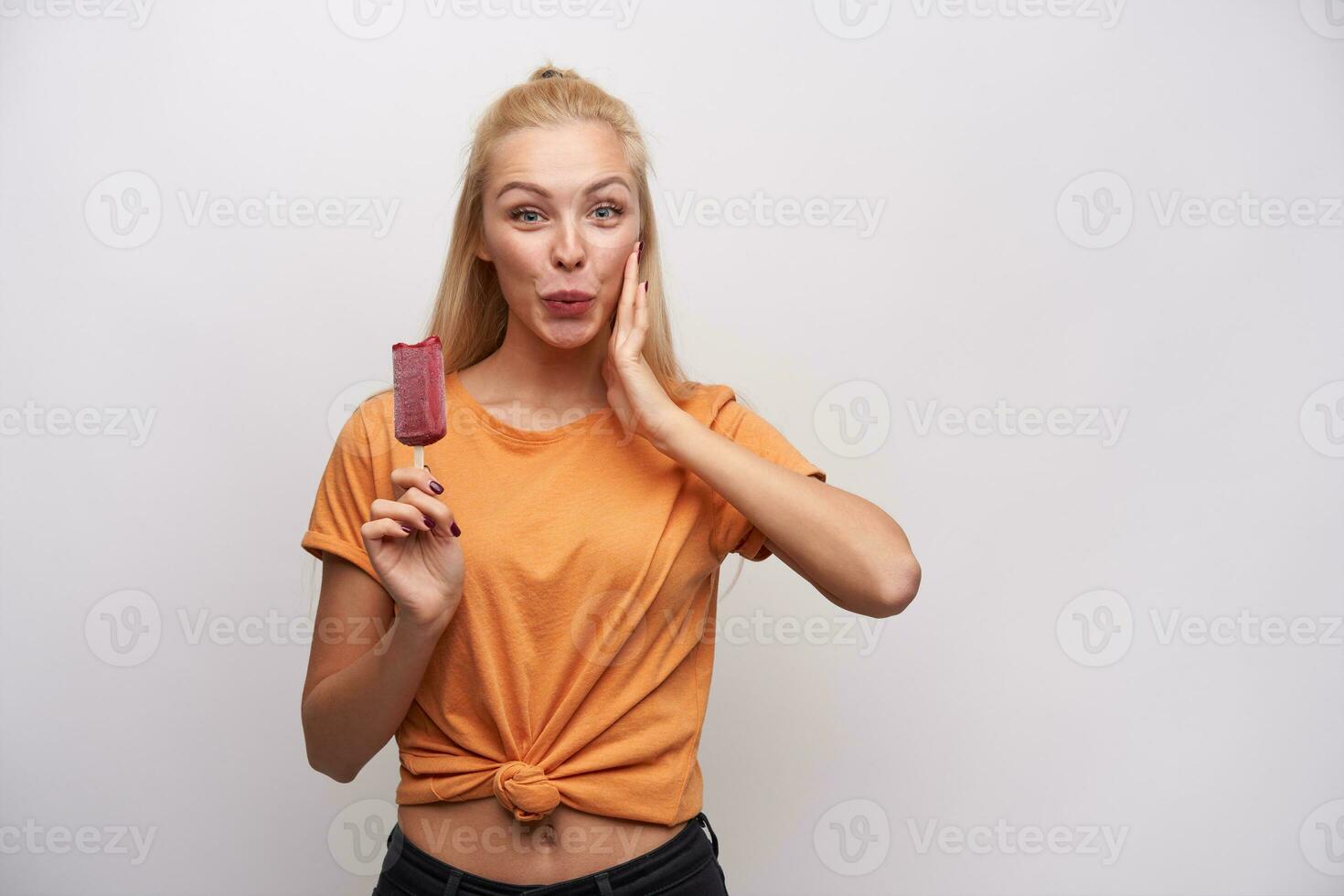 Surprised attractive young blonde lady with ponytail hairstyle looking cheerfully at camera and holding palm on her cheek, raising hand with berry ice-cream in it, standing against white background photo
