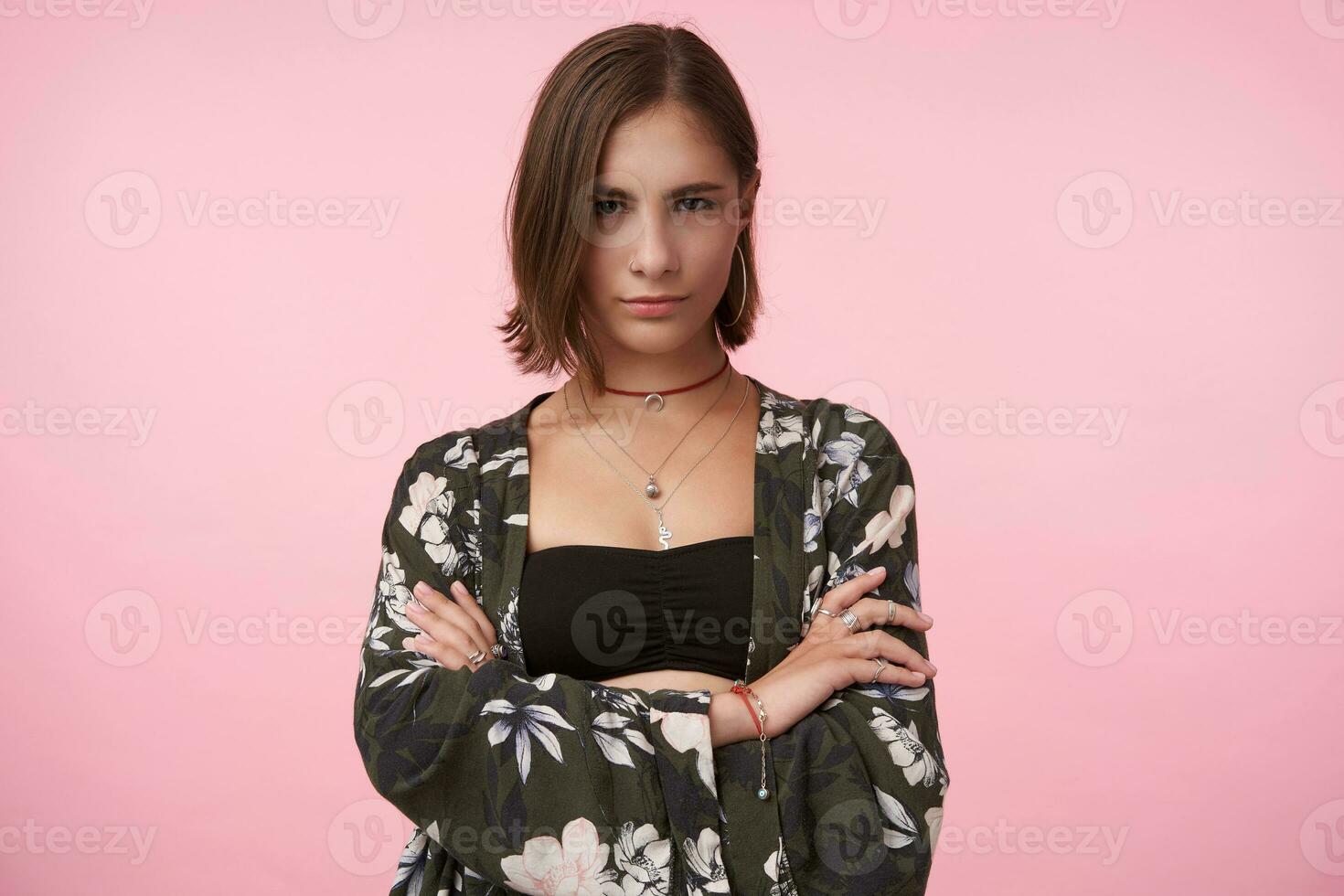 Serious young attractive brunette lady with bob haircut keeping her hands crossed while looking at camera with folded lips, standing over pink background photo