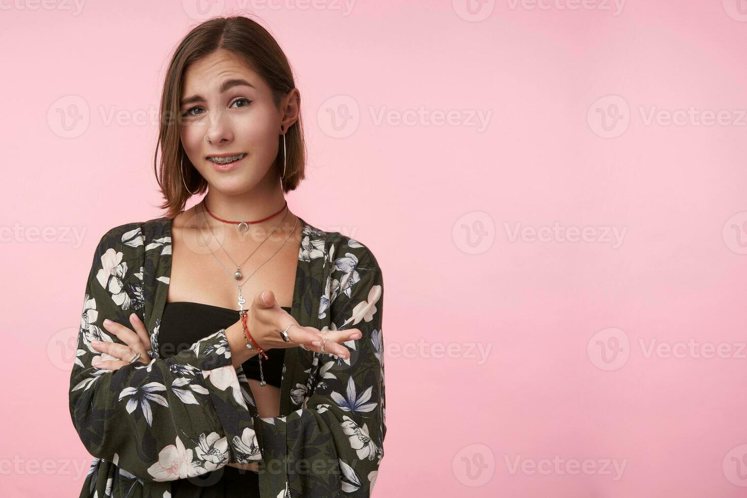 Indoor photo of young pretty brown haired woman with short haircut raising her eyebrow while looking ironically at camera, isolated over pink background