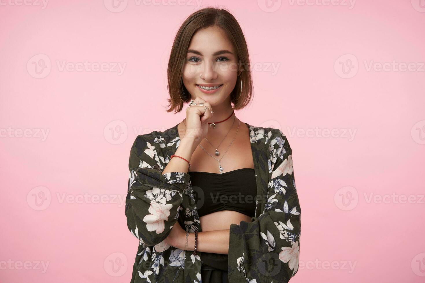 Cheerful young attractive short haired brunette female holding her chin with raised hand and smiling gladly while posing over pink background in trendy clothes photo