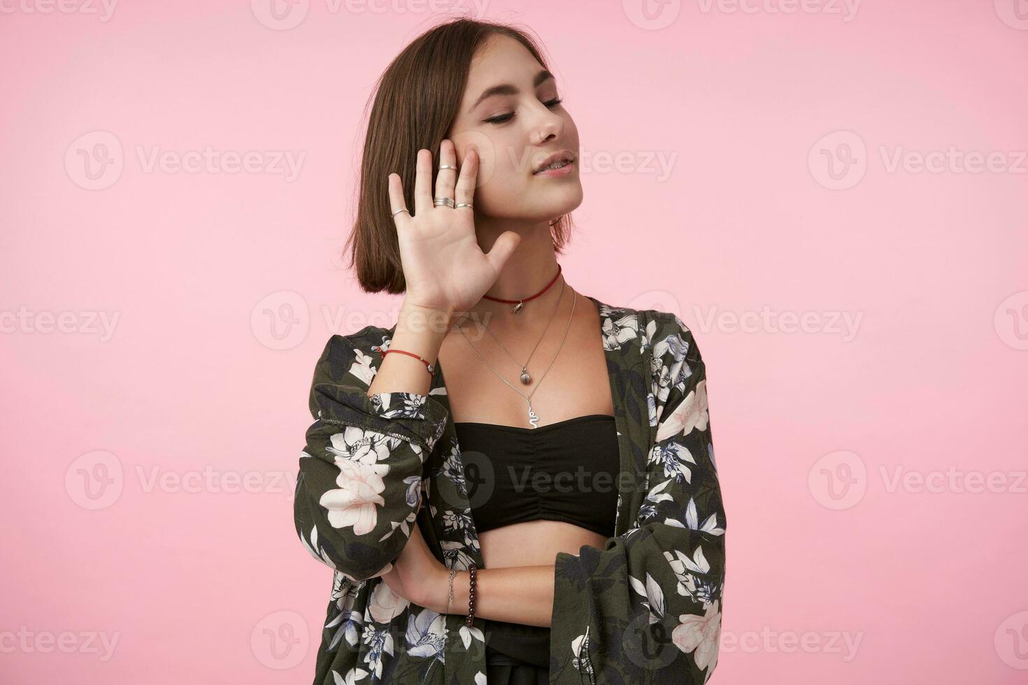 Pleasant looking young positive brown haired woman dressed in flowered shirt and black top showing raised palm and smiling slightly while posing over pink background with closed eyes photo