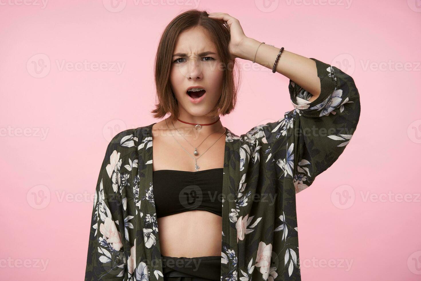 Studio photo of young puzzled short haired brunette lady frowning her eyebrows while looking confusedly at camera and keeping raised palm on her head, isolated over pink background