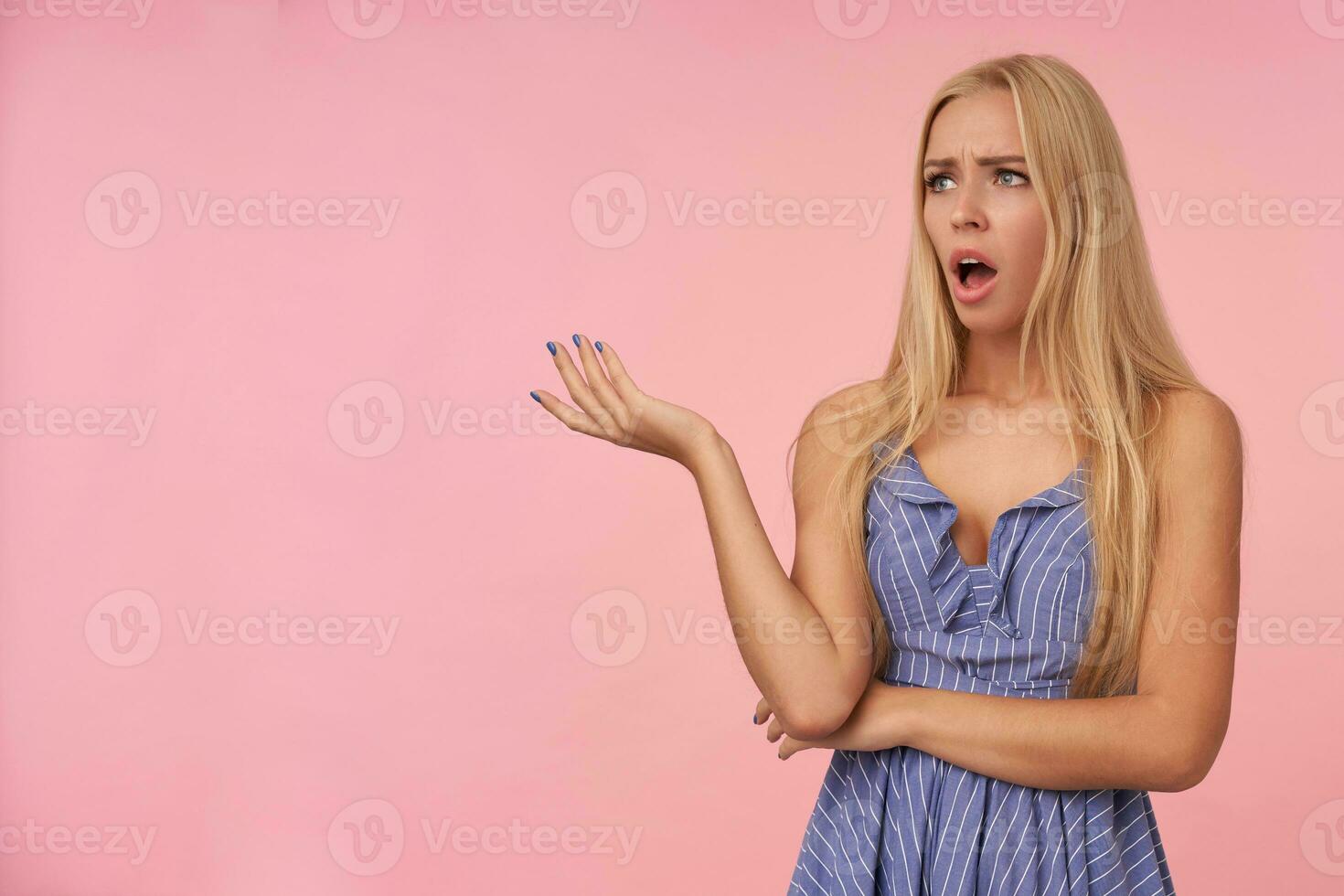Pretty bemused young blonde woman with casual hairstyle looking aside puzzled while posing over pink background, frowning with opened mouth and raising palm photo
