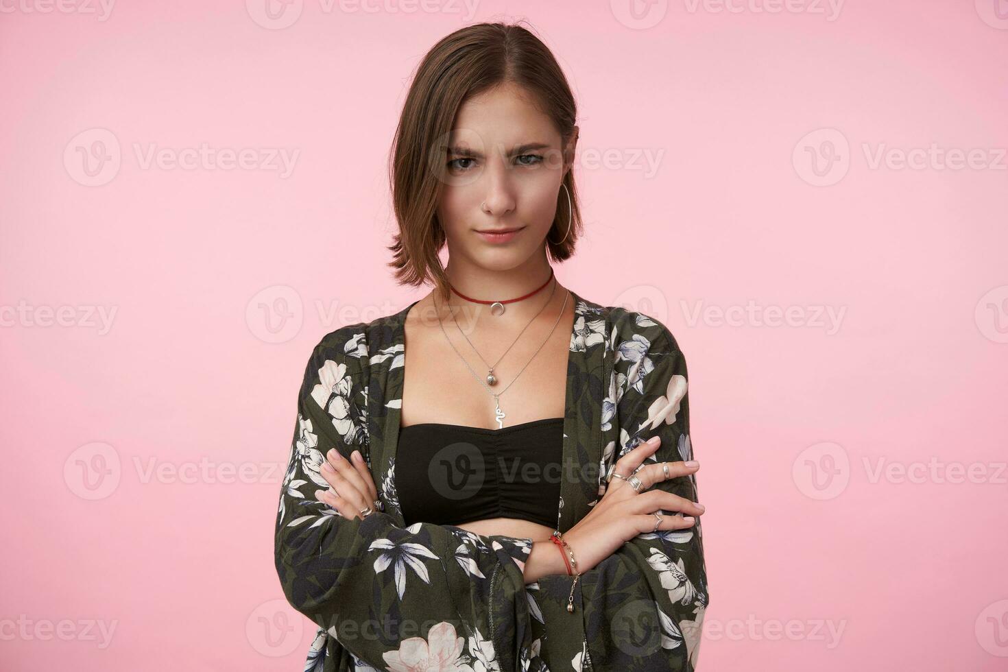 Attractive young brown-eyed brunette woman with natural makeup crossing her hands on chest while looking cunningly at camera, isolated over pink background photo