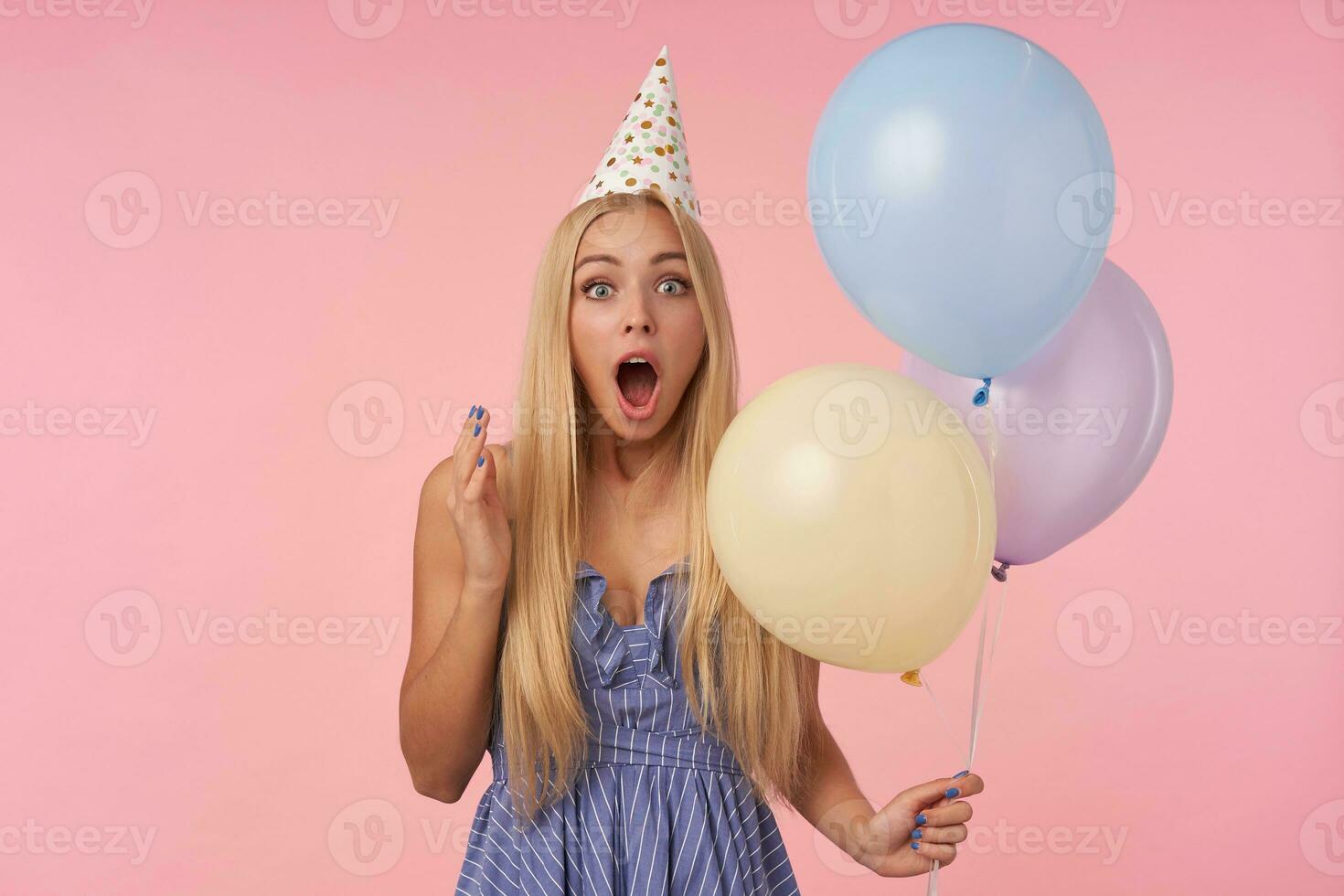 Surprised attractive long haired blonde lady in festive clothes posing in multicolored air balloons, posing over pink background with amazed face and rounding eyes photo