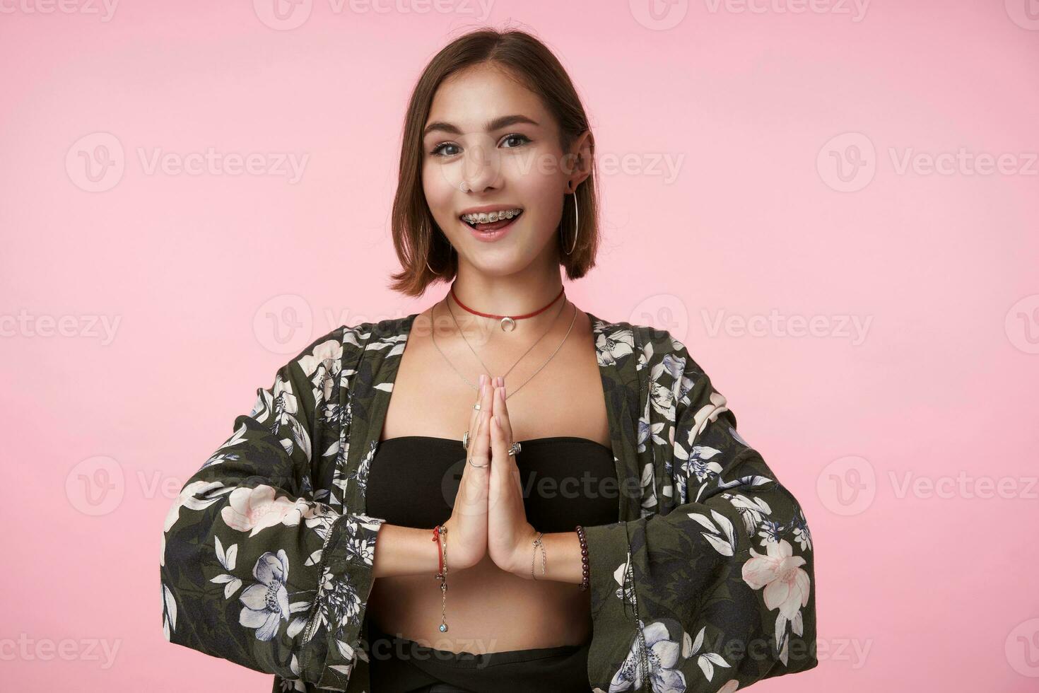 Indoor photo of young glad brown haired female with bob haircut folding raised palms together and looking excitedly at camera with broad smile, isolated over pink background