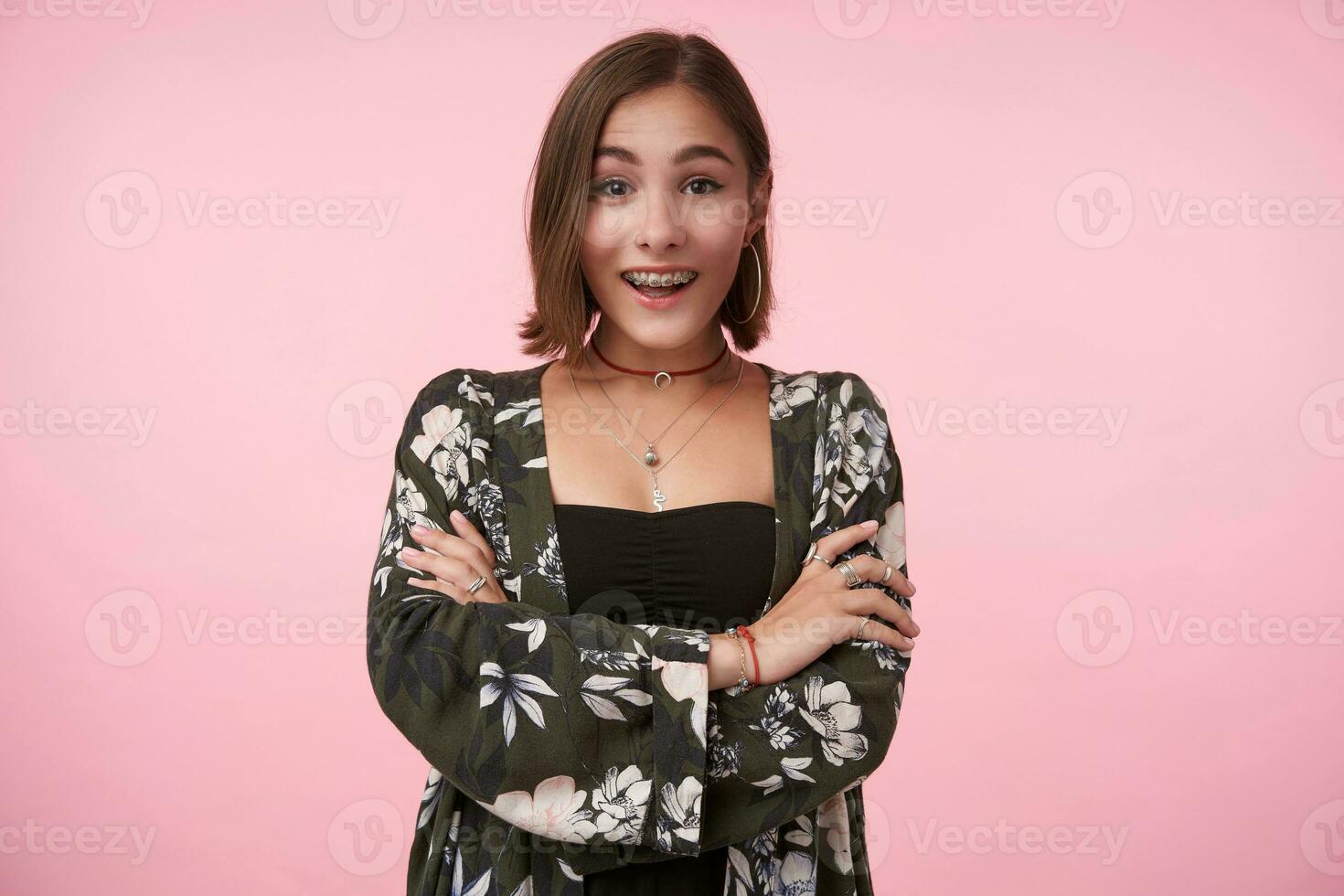 Excited young pretty short haired brunette woman folding her hands on chest while looking surprisedly at camera with wide smile, standing over pink background photo