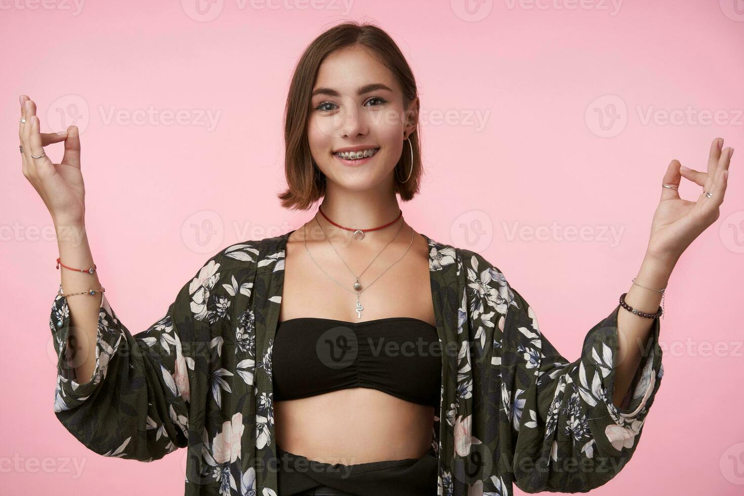 Studio shot of young cheerful short haired brown-eyed lady forming mudra sign with her fingers and smiling pleasantly at camera, standing over pink background photo