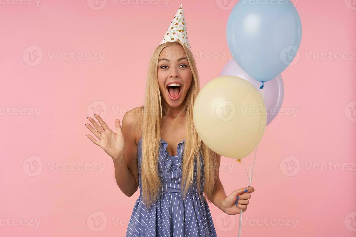 Overjoyed pretty young long haired woman with blonde hair posing in multicolored air balloons, wearing blue summer dress and birthday cap, being amused by guests while celebrating holiday photo