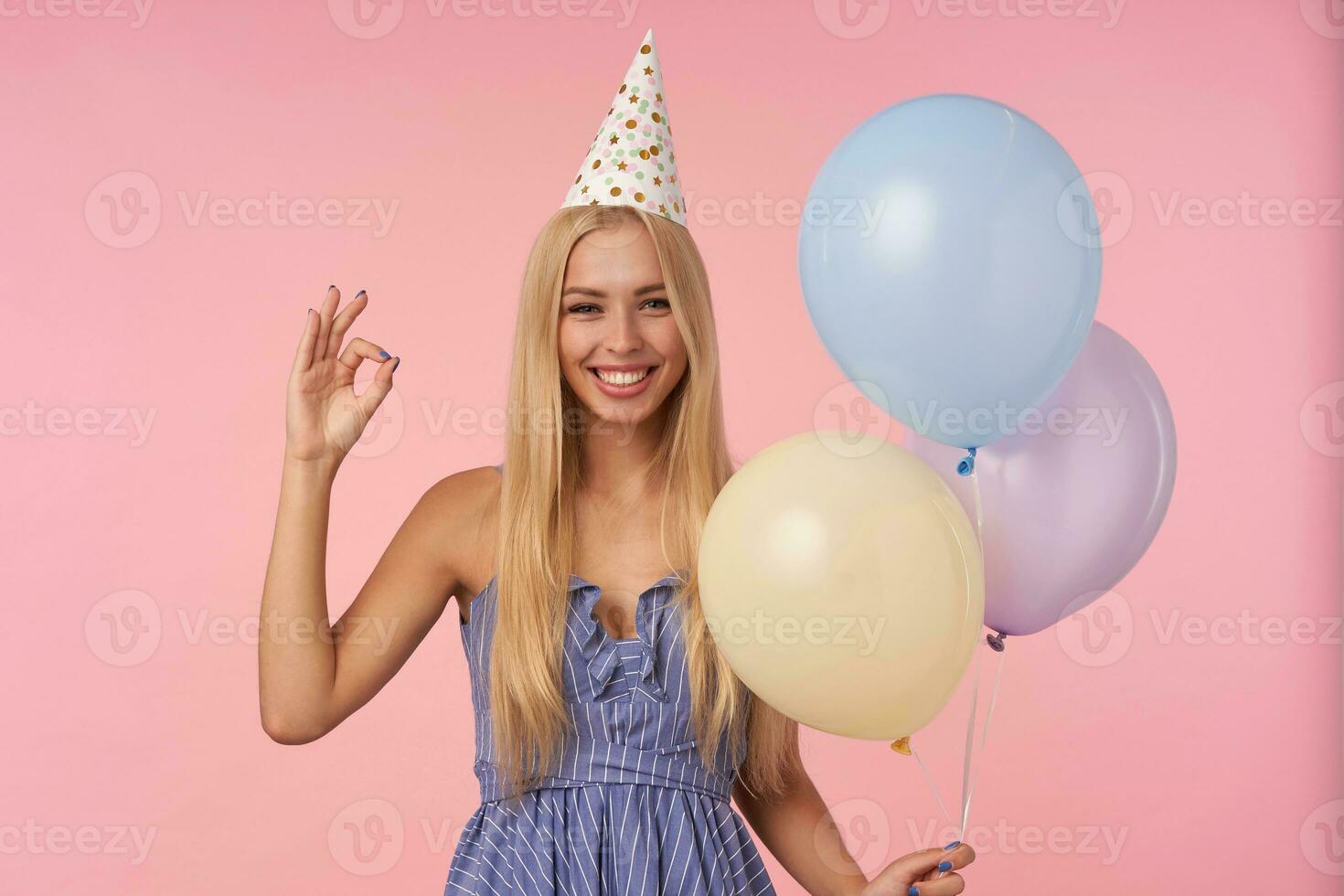 Portrait of cheerful attractive blonde female raising hand with ok gesture while standing over pink background with multicolored air balloons, looking at camera happily and smiling broadly photo