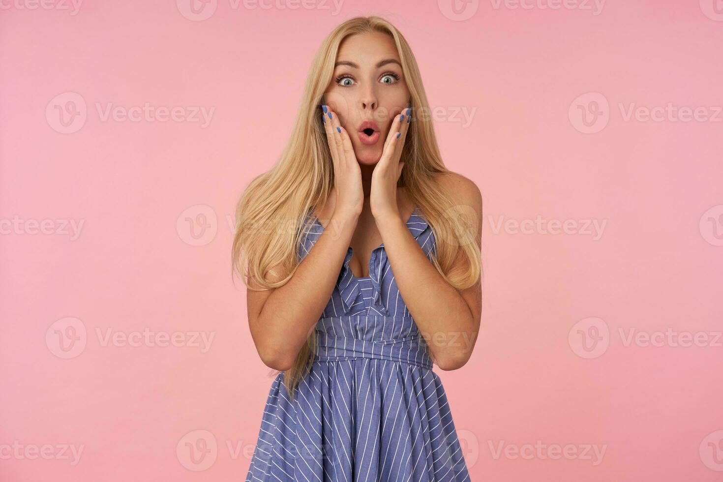 Portrait of astonished young long haired blonde lady looking at camera with folded lips and keeping palms on her cheeks, posing over pink background in casual clothes photo