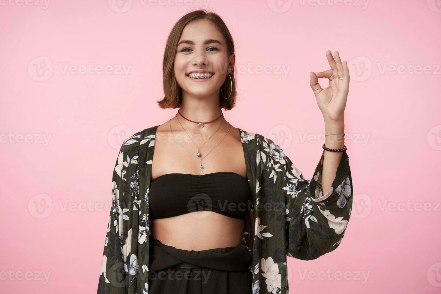Happy young attractive brunette lady dressed in flowered shirt and black top showing well done sign and smiling cheerfully while posing over pink background photo