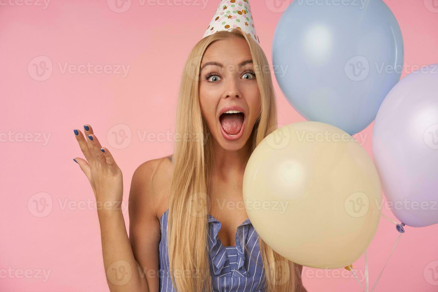 Joyous young attractive woman with long blonde hair casual hairstyle showing happy reaction on getting awesome present, posing in multicolored air balloons while standing over pink background photo