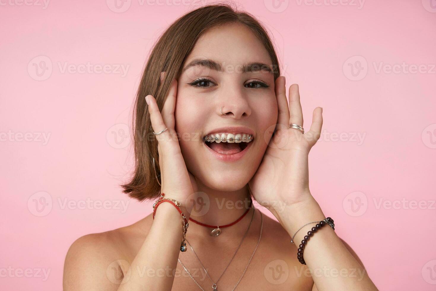 Cheerful young lovely brown-eyed brunette woman with short haircut holding her face with raised hands and smiling happily while posing over pink background photo