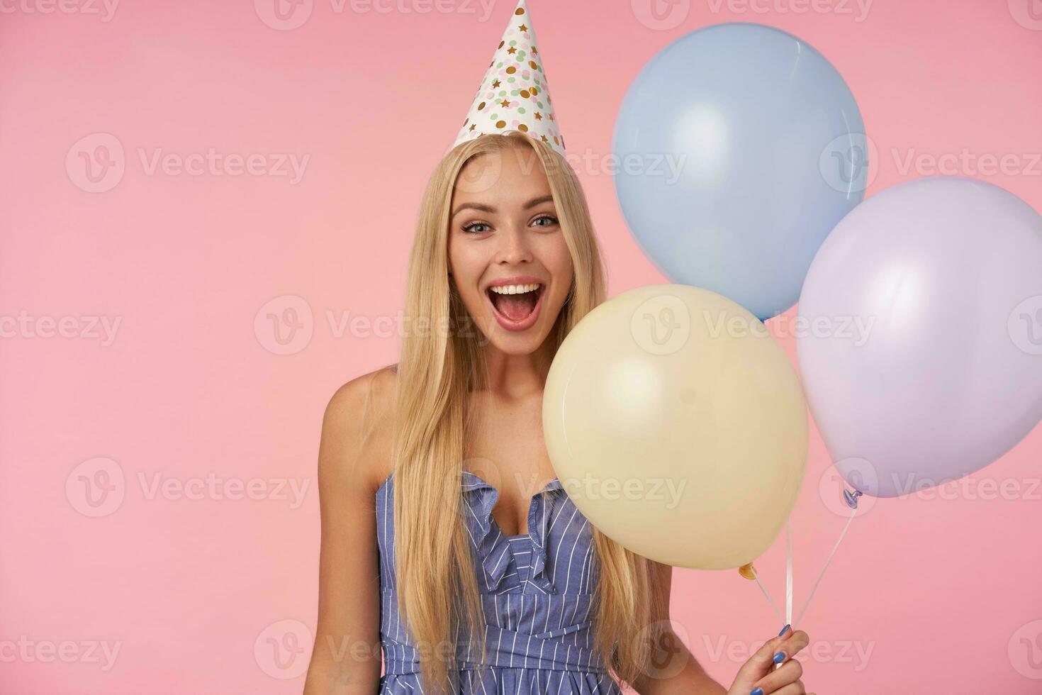 Joyful pretty long haired blonde lady having cheerful moments in her life during birthday party, posing over pink background with multicolored air balloons, being in high spirit photo