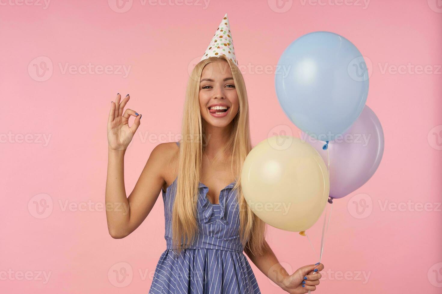 Joyous long haired blonde lady in festive clothes rejoicing while posing in multicolored air balloons, isolated over pink background with folded fingers in ok sign,smiling at camera and showing tongue photo