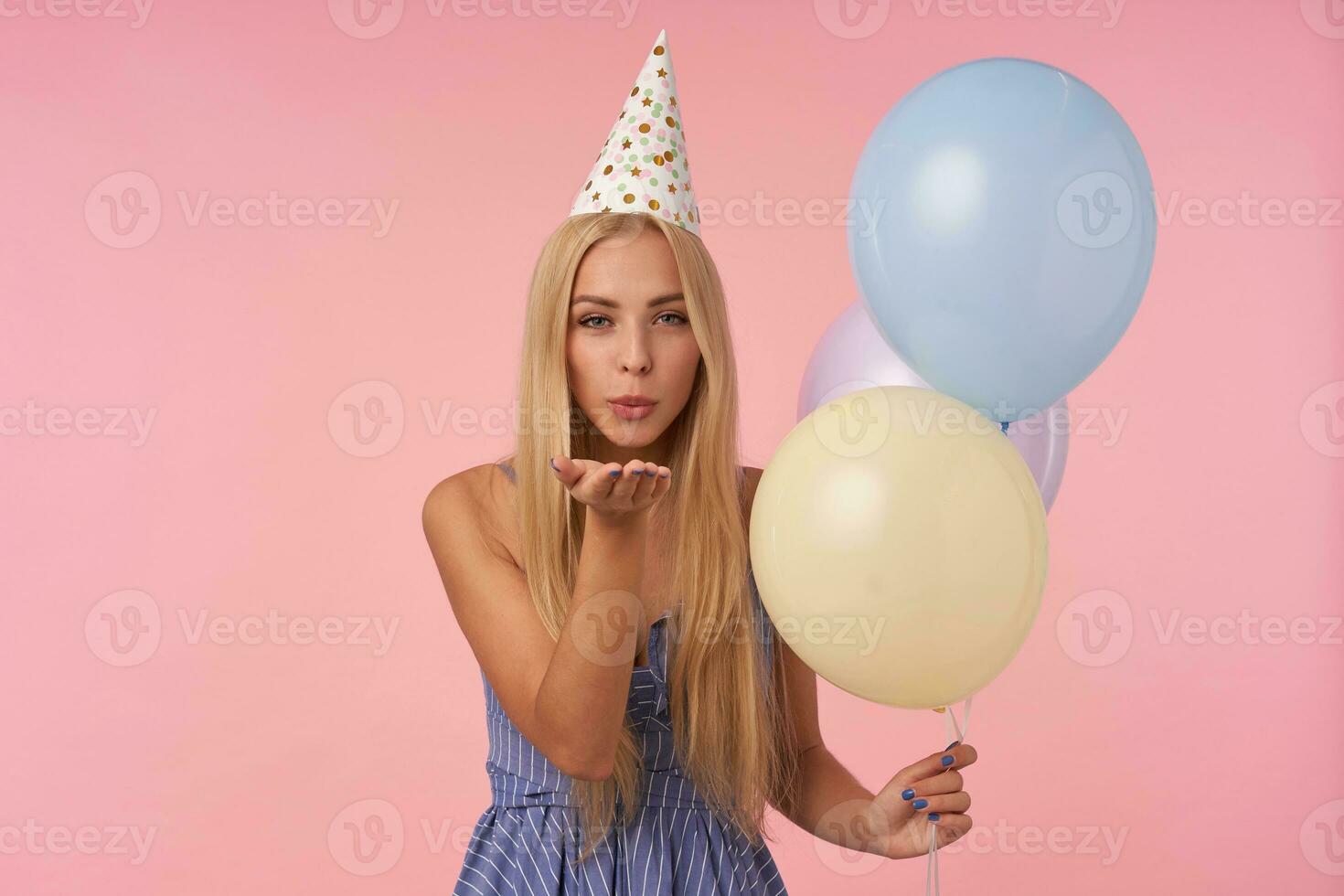 Portrait of attractive young long haired woman celebrating birthday with multicolored air balloons, looking at camera positively and blowing air kiss with pursed lips photo