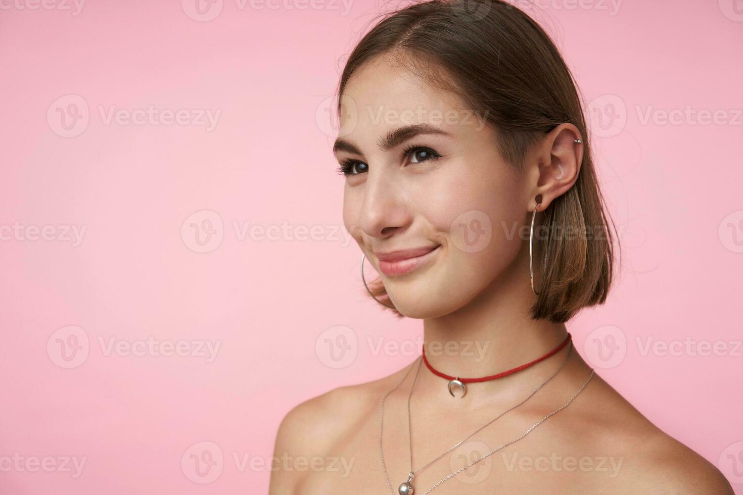 interior foto de joven encantador corto peludo morena hembra sonriente agradablemente mientras mirando afirmativamente aparte, en pie terminado rosado antecedentes con desnudo espalda