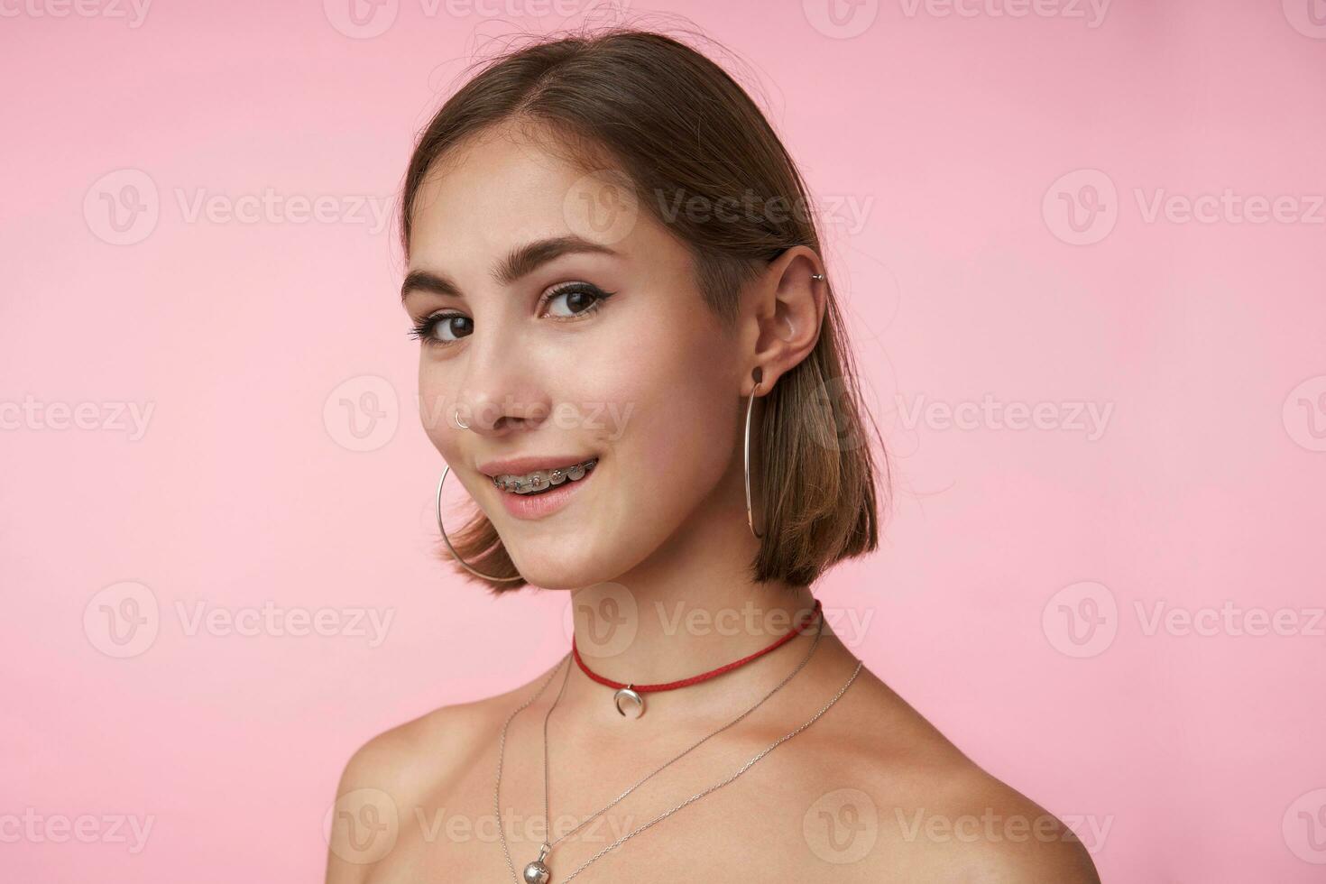 Close-up of young pretty brown-eyed brunette woman with natural makeup smiling cheerfully while looking at camera, wearing trendy accessories while posing over pink background photo