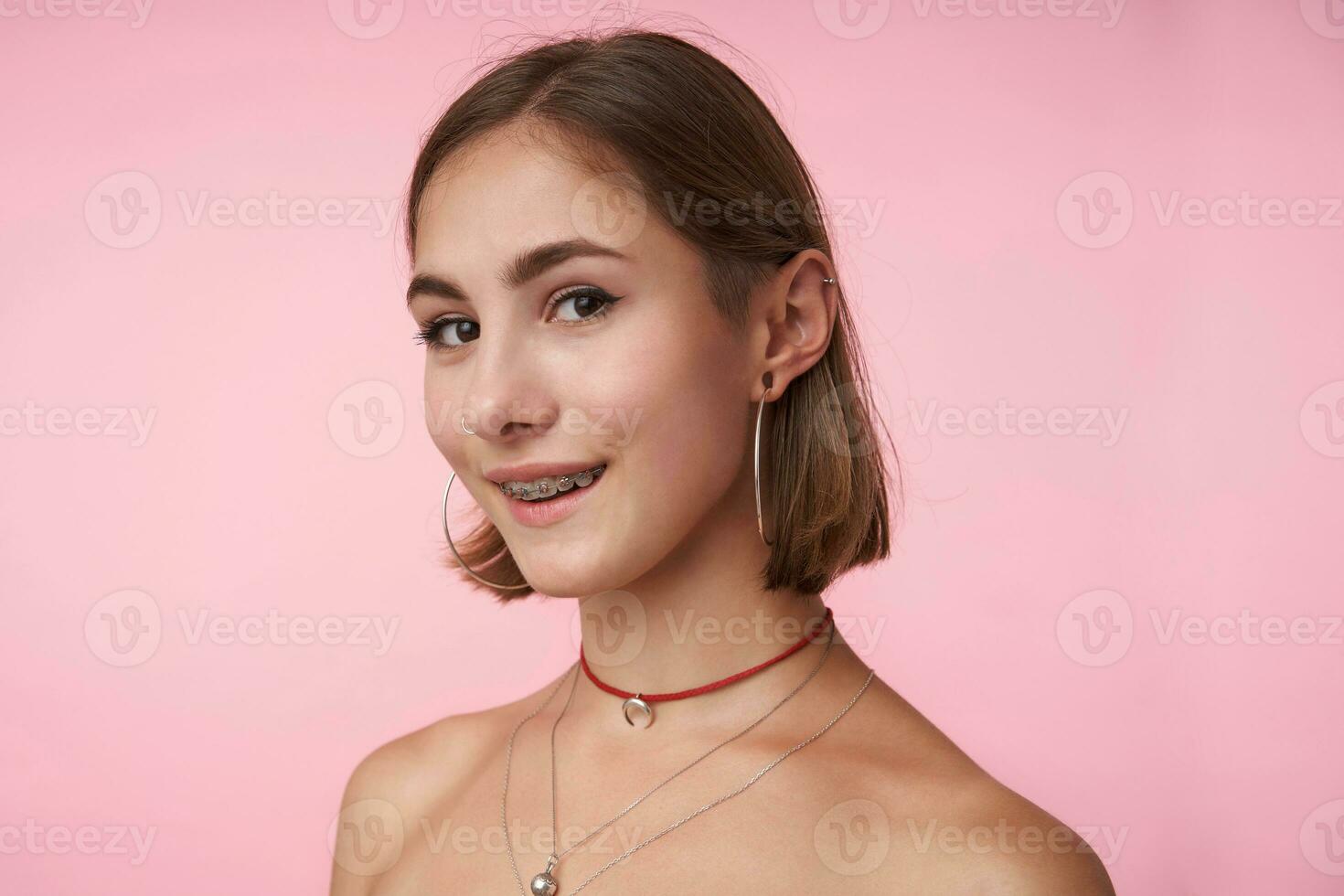 de cerca de joven bonito de ojos marrones morena mujer con natural maquillaje sonriente alegremente mientras mirando a cámara, vistiendo de moda accesorios mientras posando terminado rosado antecedentes foto