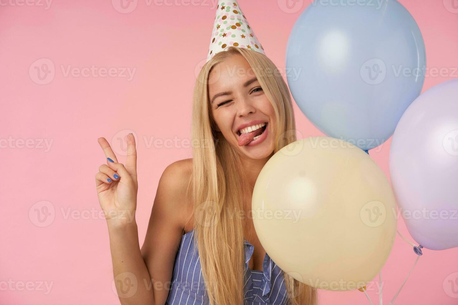 Funny shot of long haired blonde female in blue summer dress and birthday cap standing over pink background, winking at camera with sticking out tongue and raising vistory gesture photo