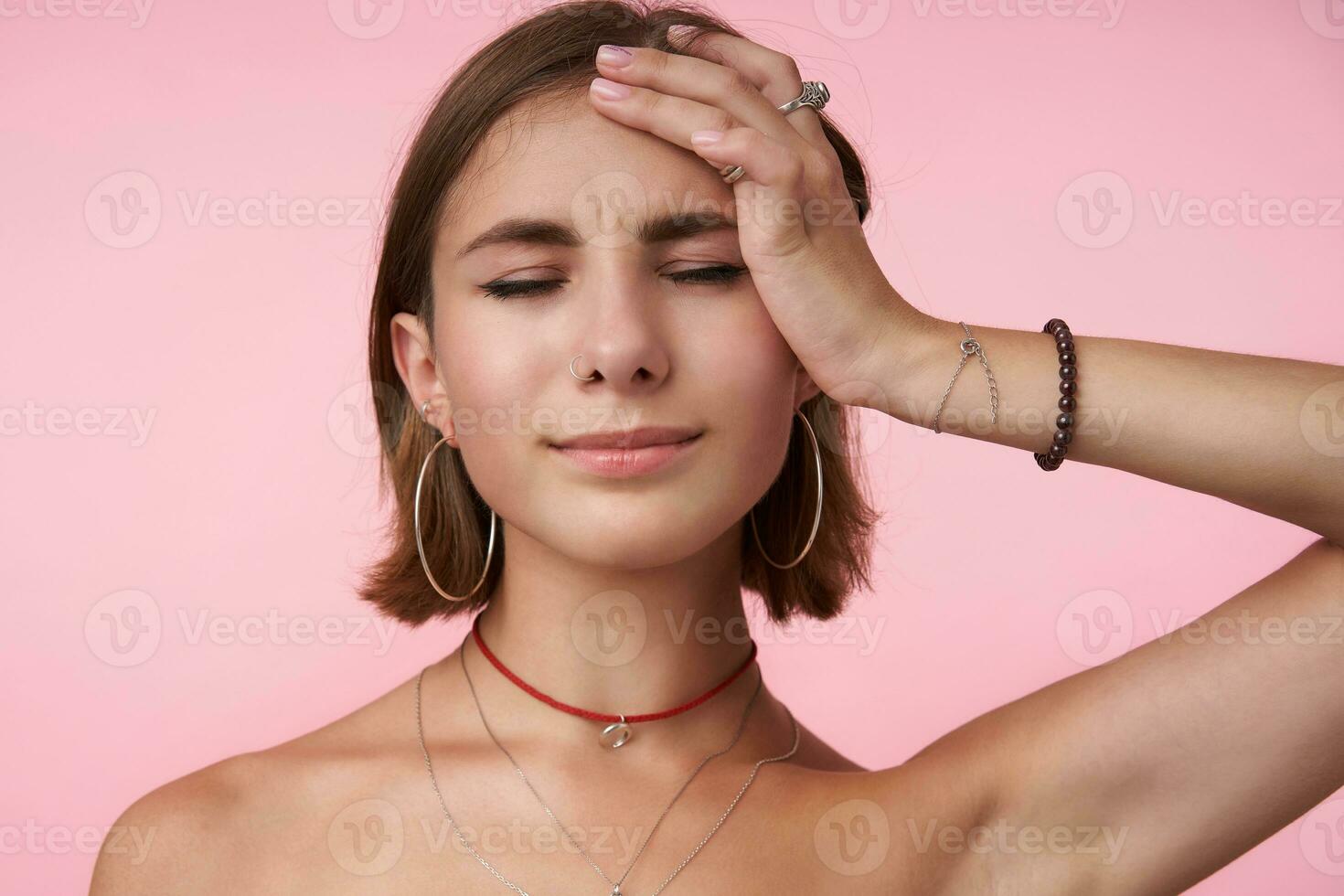 Studio shot of young good looking brunette female frowning her face and keeping raised palm on her head while standing over pink background with closed eyes photo
