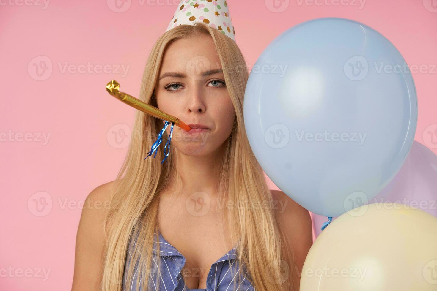 Uninspired young long haired blonde female posing indoor with multicolored helium balloons, blowing party horn and looking at camera with bored face, standing over pink background photo