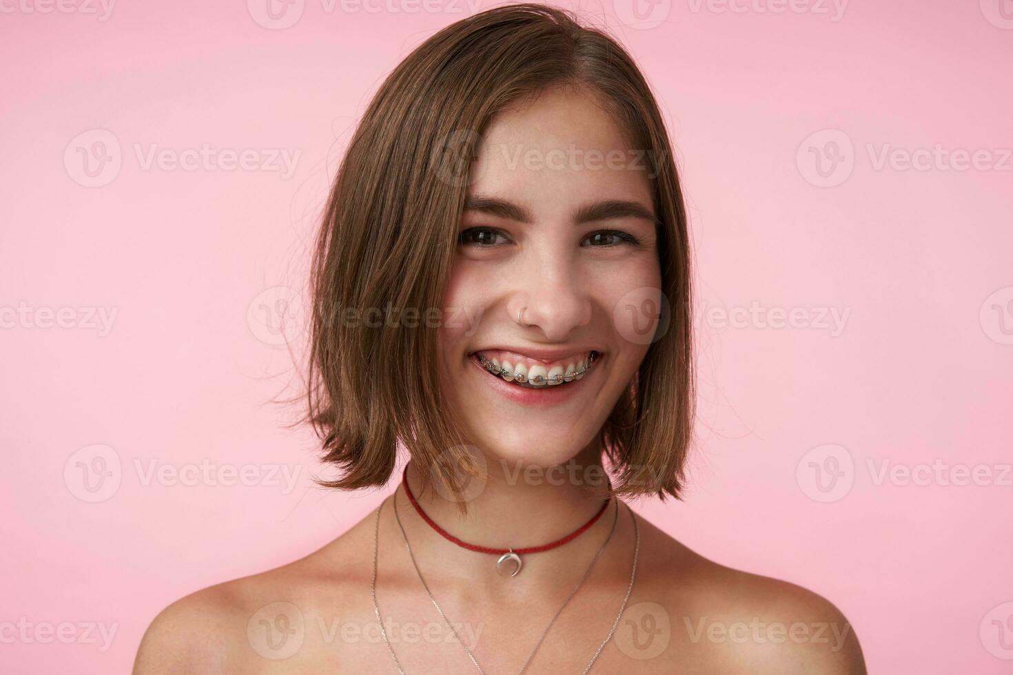 Close-up of young cheerful brown-eyed brunette female with bob haircut looking happily at camera with charming smile, isolated over pink background photo