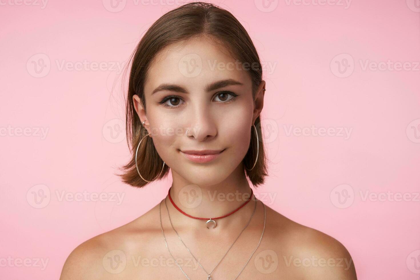 horizontal foto de joven de ojos marrones morena mujer mirando afirmativamente a cámara y sonriente levemente, en pie terminado rosado antecedentes con manos abajo