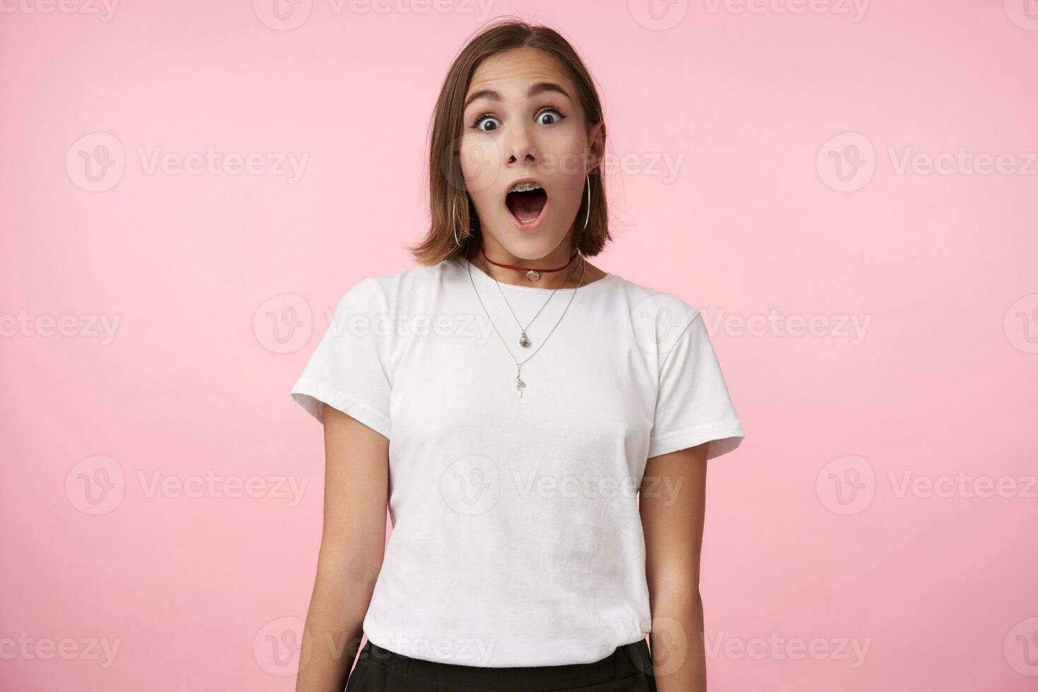 Shocked young pretty brown haired woman dressed in casual clothes looking excitedly at camera with wide eyes and mouth opened, posing over pink background photo