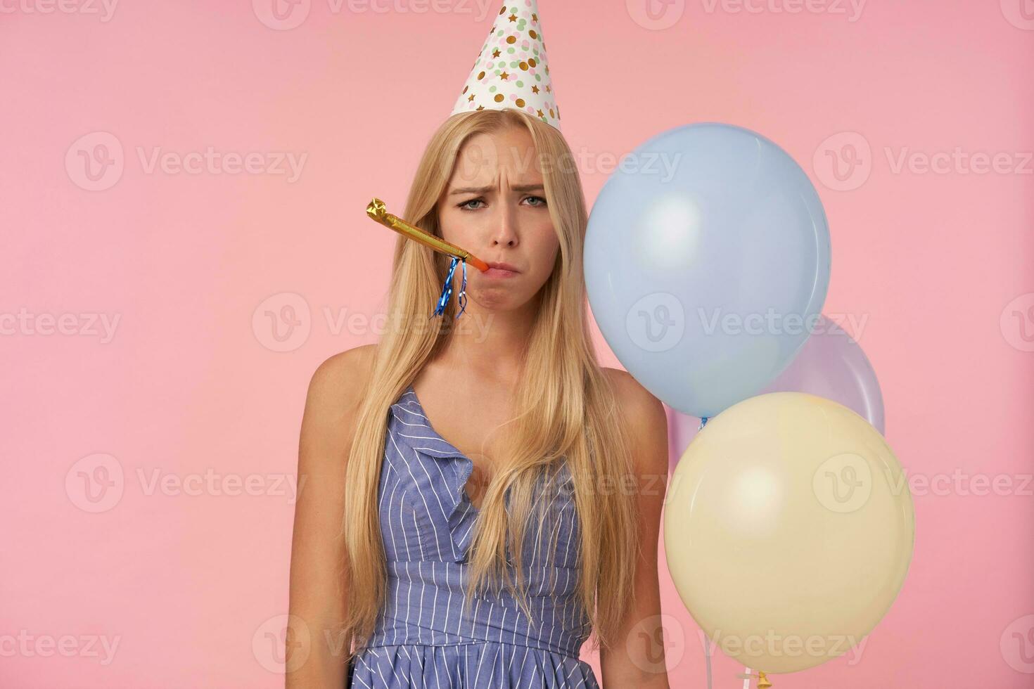Photo of unpleased long haired blonde lady in blue summer dress and holiday cap blowing party horn casual over pink background, looking at camera with upset face and holding bunch of helium balloons