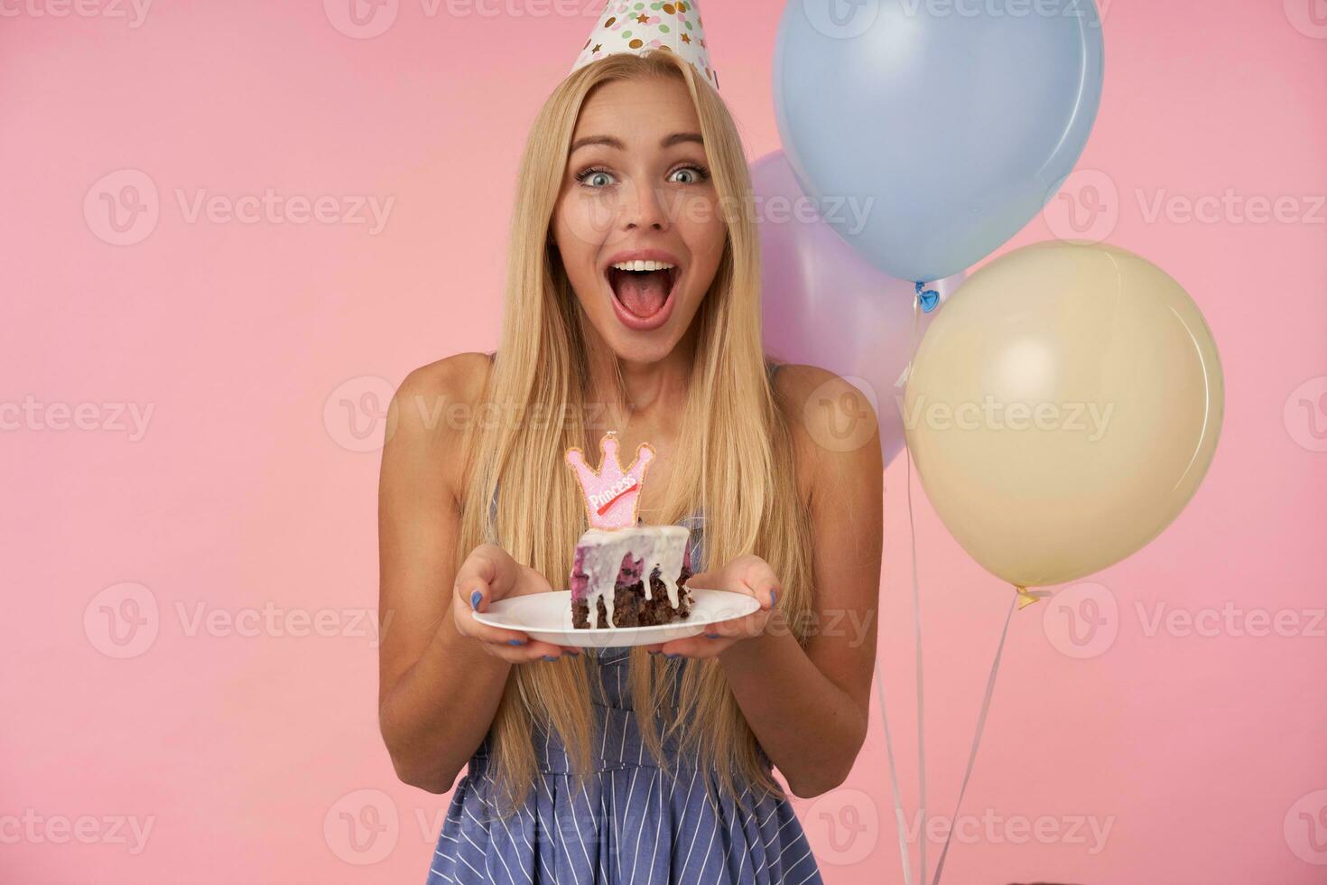 Joyful young pretty woman with long blonde hair wearing blue summer dress and cone hat, celebrating birthday and keeping piece of cake in hands, smiling widely over pink background photo