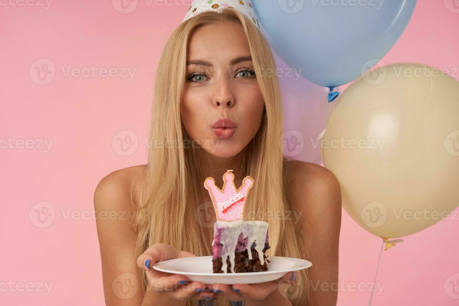 Delighted young blonde woman looking at camera and folding lips to blow on candle, showing her pleasant emotions while posing over pink background with helium multicolored balloons photo