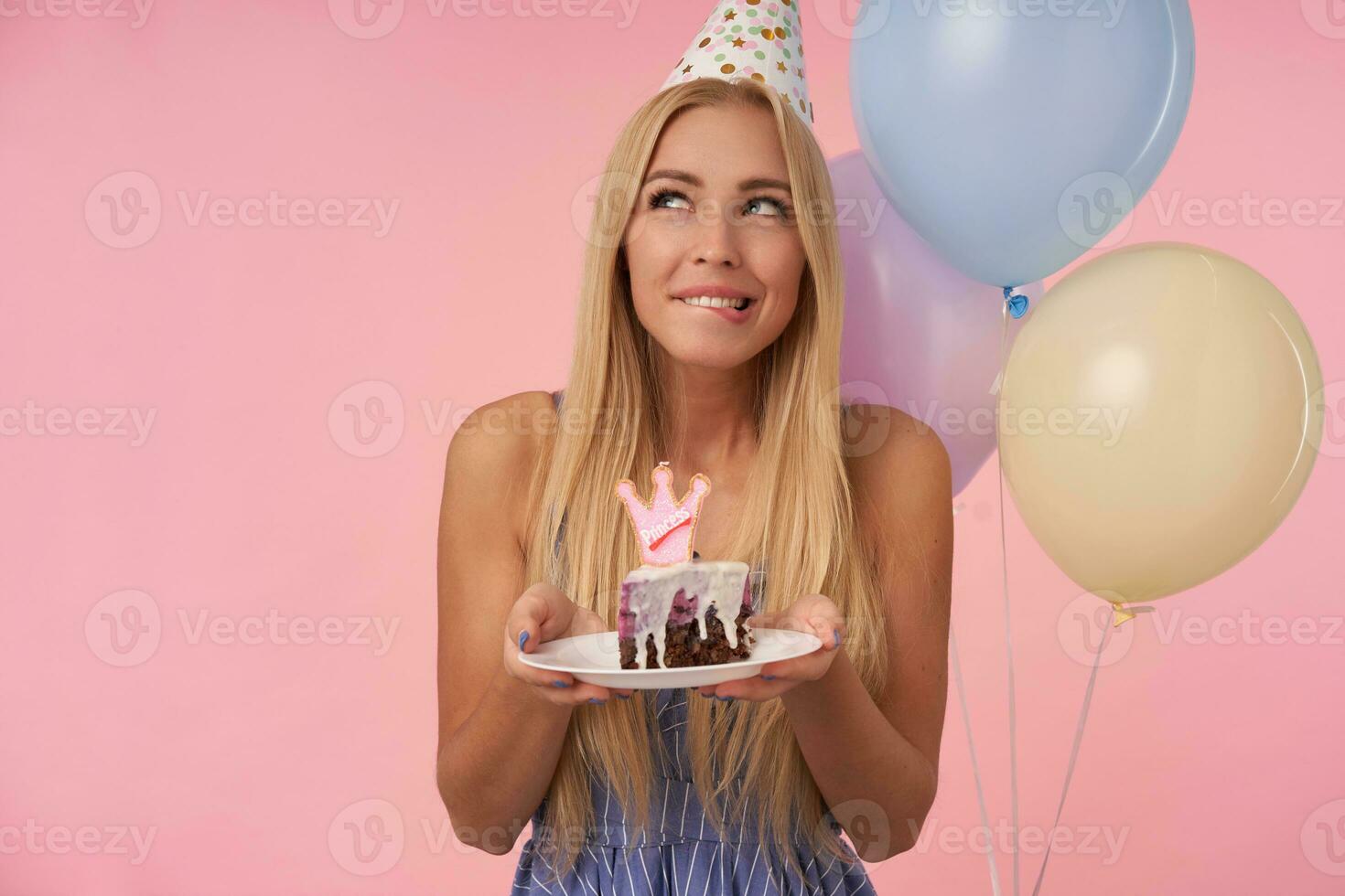 Indoor shot of attractive long haired blonde lady rejoicing while posing in multicolored air balloons, holding piace of cake and looking aside thoughtfully, biting underlip over pink background photo