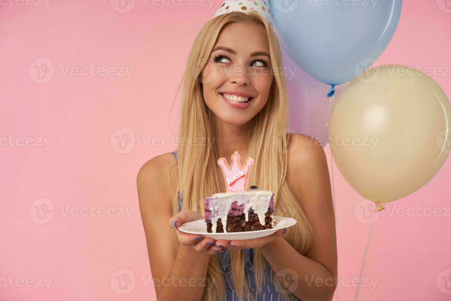 Attractive young blonde lady with piece of cake in her hands looking aside dreamily with wide happy smile, wearing blue summer dress and cone hat while standing over pink background photo