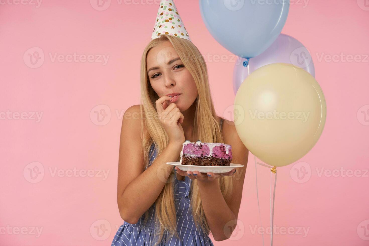Beautiful pleased long haired female in blue summer dress and holiday cone hat tasting yummy pie over pink background, licking her forefinger and looking at camera with happy smile photo