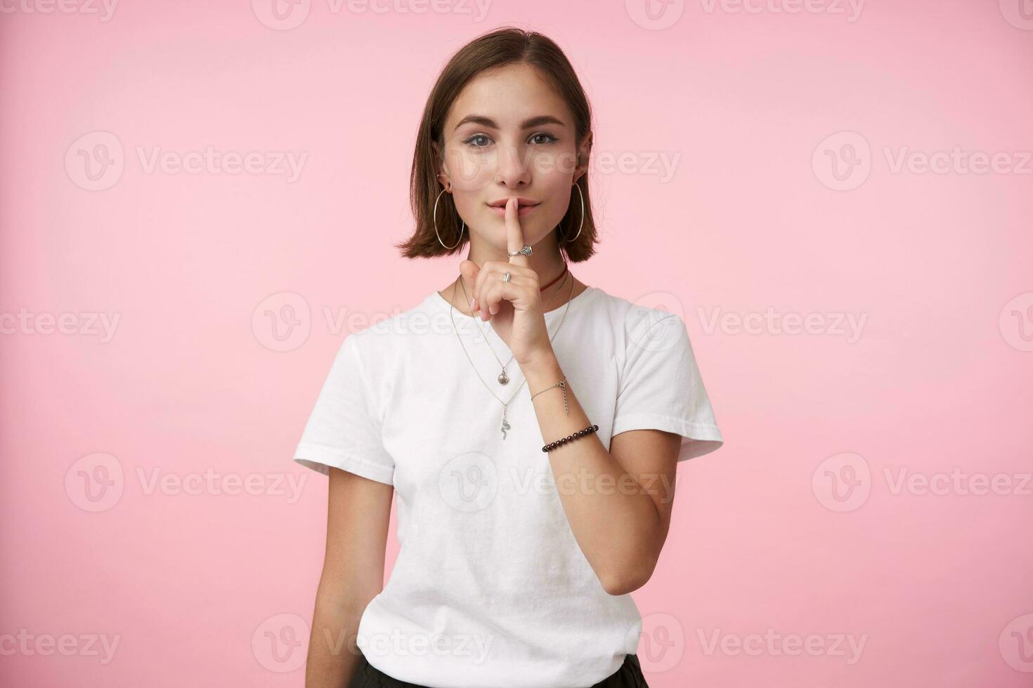 Positive young brown-eyed brunette woman with natural makeup raising hand with hush gesture and holding index finger on her mouth, isolated over pink background photo