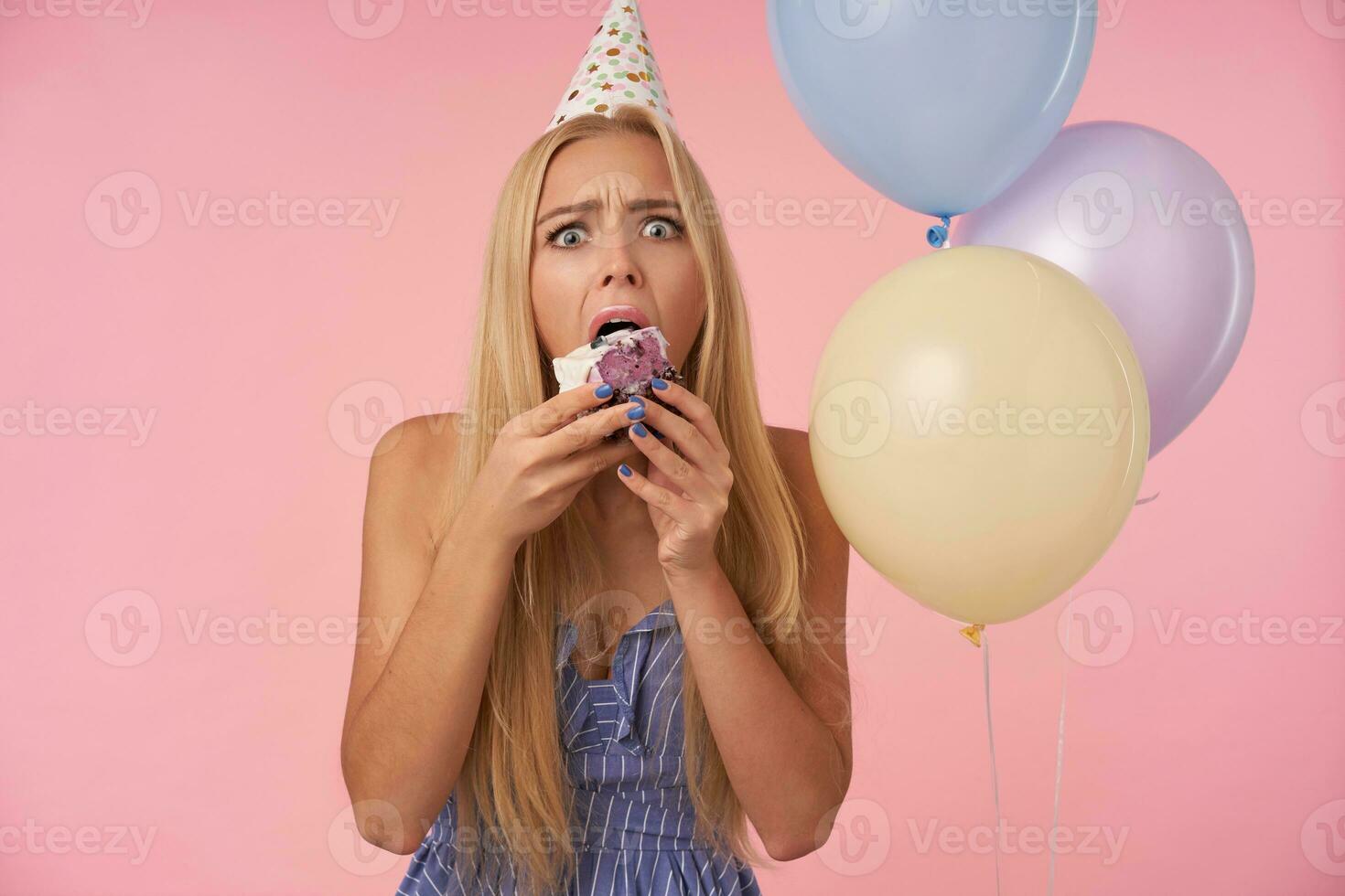 Indoor photo of young pretty female with long blonde hair wearing blue summer dress and birthday cap, being surprised while eating cake and looking at camera with wide eyes opened