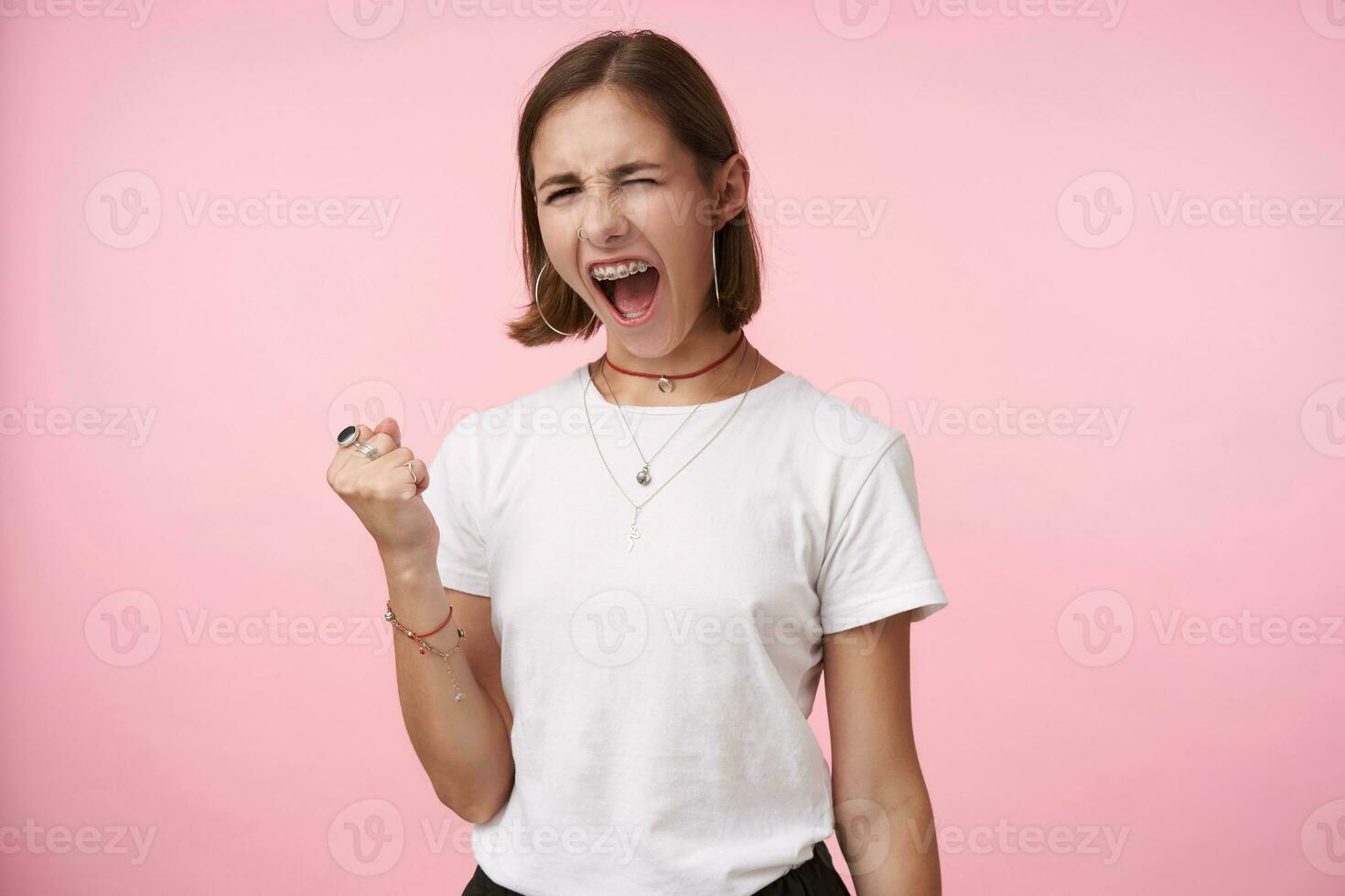 Indoor photo of expressive young brown haired woman with natural makeup keeping her mouth wide opened while screaming excitedly, isolated over pink background