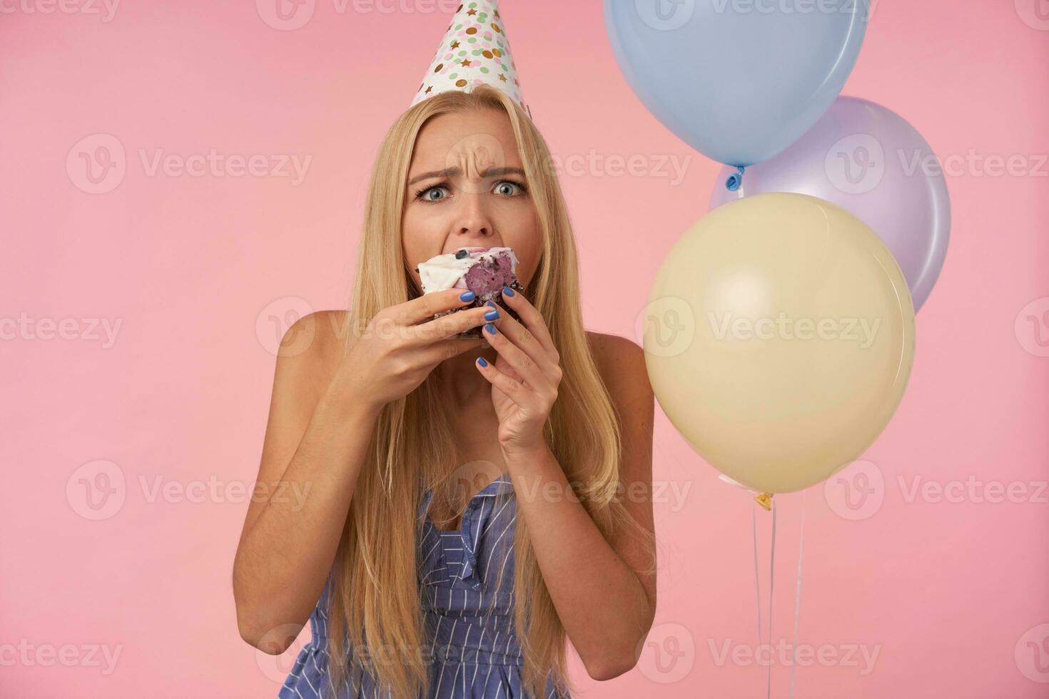 Amazed young blonde woman in festive clothes and cone hat frowning her eyebrows and rounding eyes while eating delicious pie in colored helium balloons, isolated over pink background photo